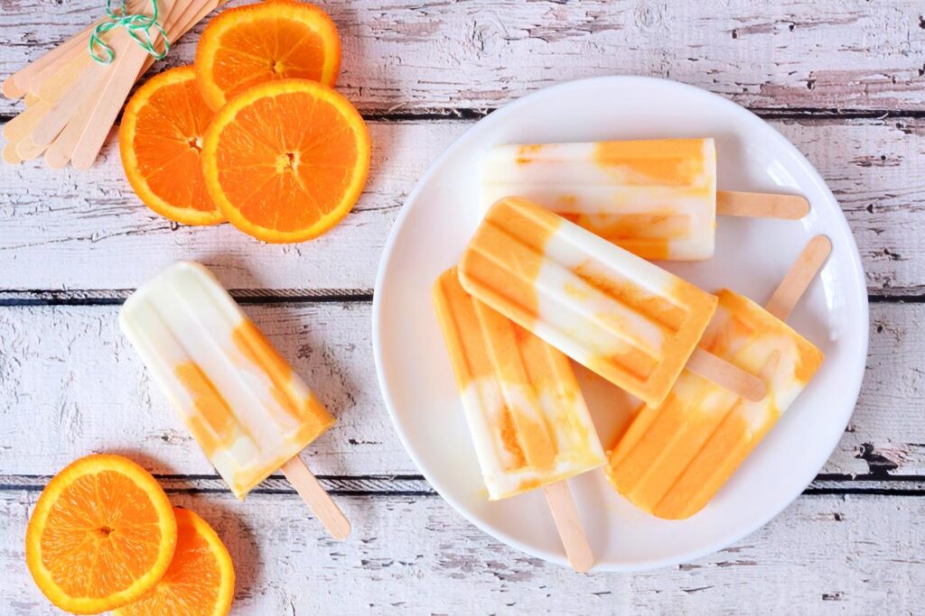 Delicious homemade citrus popsicles on a plate next to orange slices all on a rustic looking white picnic table