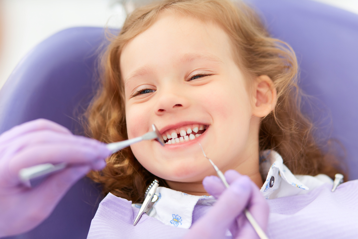 A smiling little girl with auburn colored hair smiling while her teeth are being examined by a dental hygienist or dentist wearing purple colored gloves on their hands 