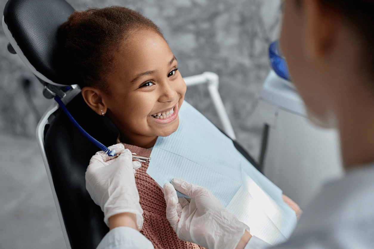 A school-age African American girl in a dental chair. She is smiling as a dental technician places a protective bib on her before her treatment starts. 