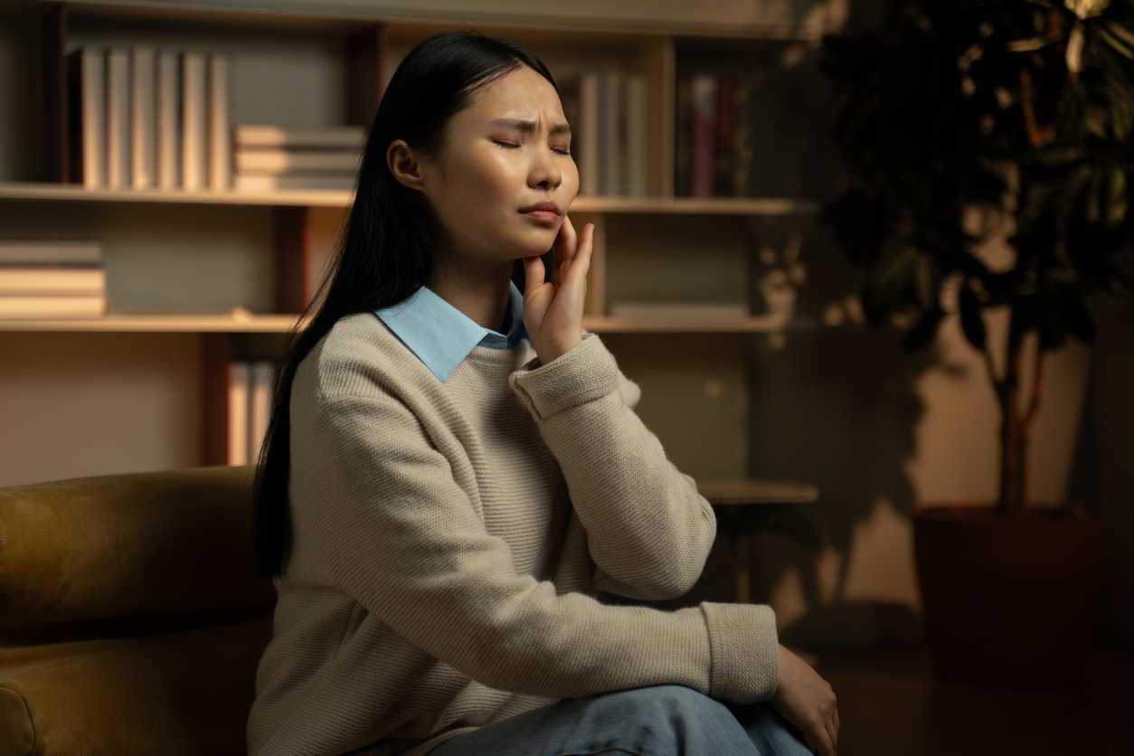 A young Asian woman suffering dental pain in her home. She has her left hand resting against her left jaw, and her eyes are closed in pain.
