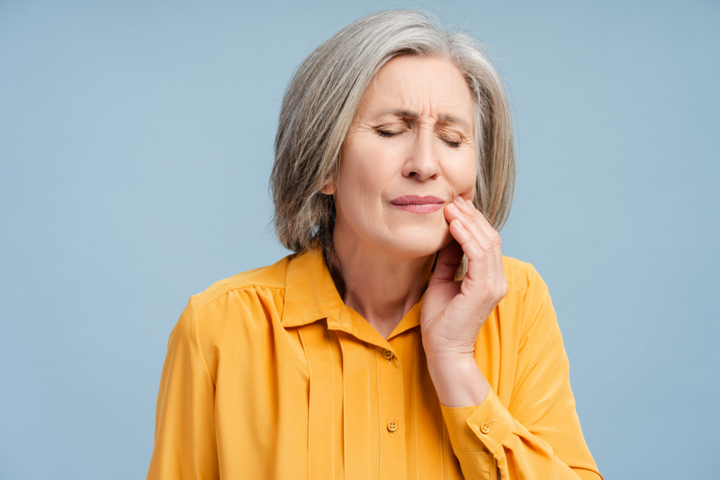 Frustrated older woman suffering from dental pain, holding her left hand to her cheek. 