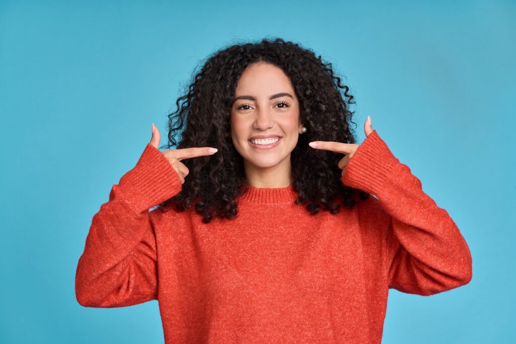Young dark-haired woman smiling broadly and pointing to her healthy teeth and gums.
